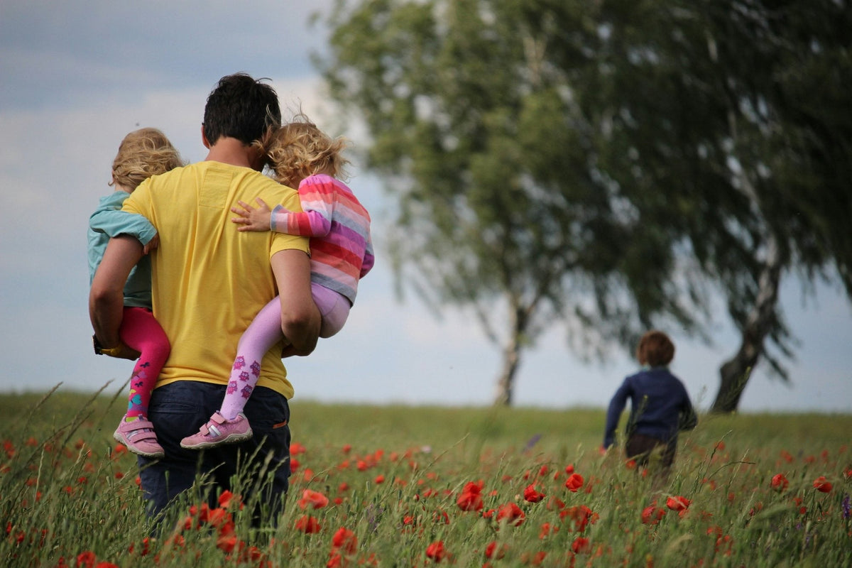 A father carrying his two young daughters through a summer poppy meadow while an older child runs ahead